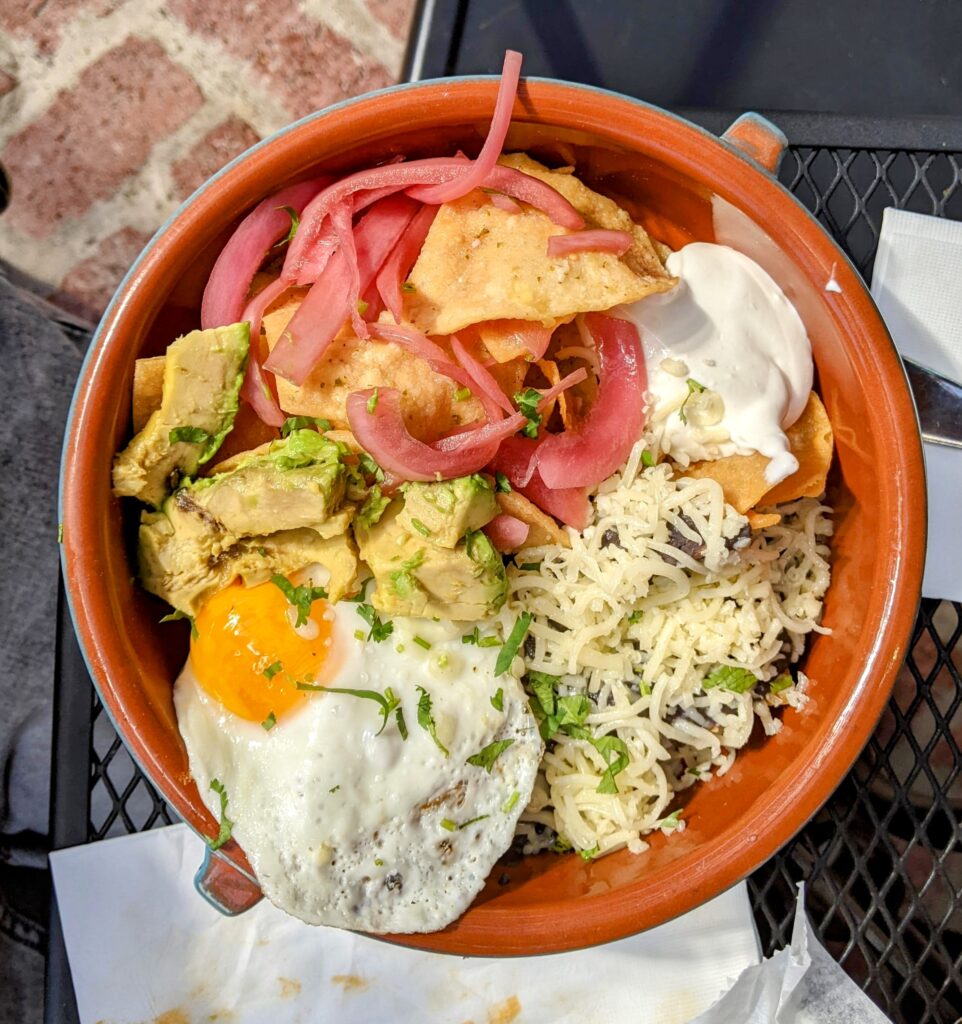 A beautiful terracotta-colored bowl with an egg, avocado wedges, shredded cheese, sour cream, chips, and pickled red onion.